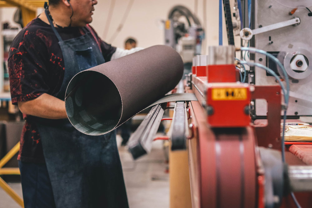 Worker carrying a wide sanding belt beside an industrial belt-making machine