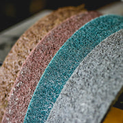 Stack of 25 x 48 surface conditioning belts in brown, maroon, blue, and grey for various grit finishes on metal surfaces.